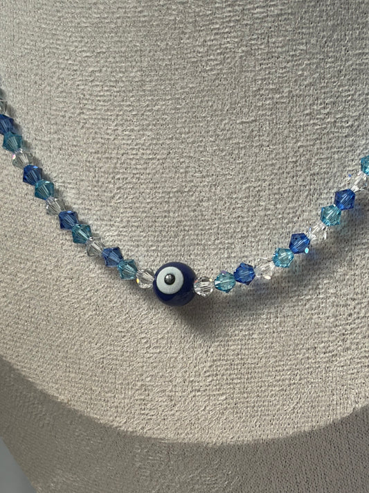 A Swarovski crystal beaded necklace with blue and light blue faceted beads and a central evil eye charm, displayed on a textured beige jewelry bust.