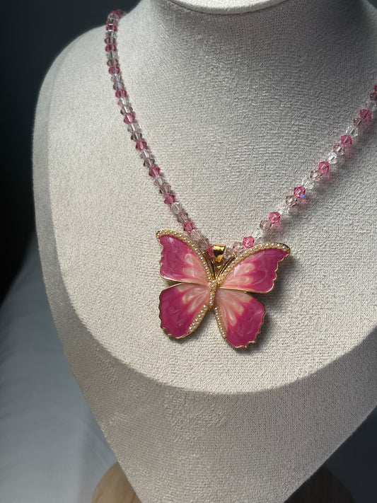 A pink butterfly pendant necklace with gold-tone detailing and a Swarovski crystal beaded chain, displayed on a textured light gray jewelry bust.