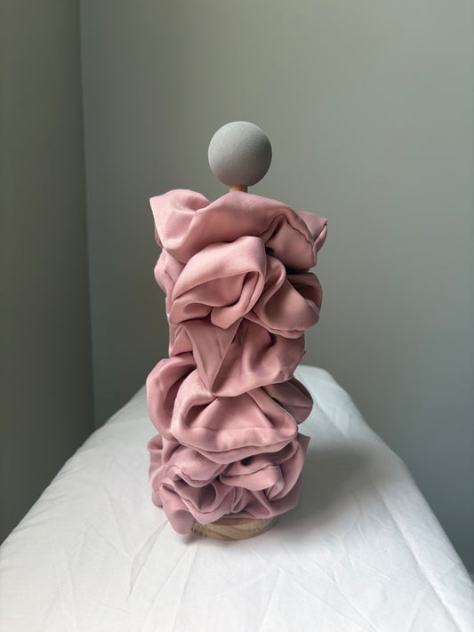A sculptural stick of scrunchies from layered dusty pink fabric ruffles, topped with a round gray sphere on a wooden stand, set on a white draped surface against a gray wall.