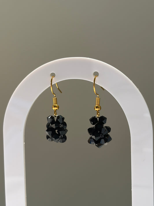 A pair of gold-tone dangle earrings with faceted black Swarovski crystal beaded clusters, displayed on a white arched stand against a neutral gray background.