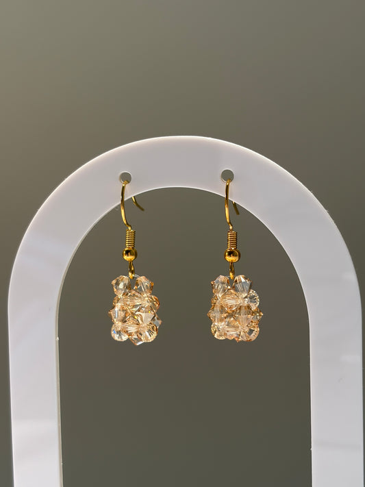 A pair of gold-tone dangle earrings with faceted gold-toned Swarovski crystal beaded pendants, displayed on a white arched stand against a neutral gray background.