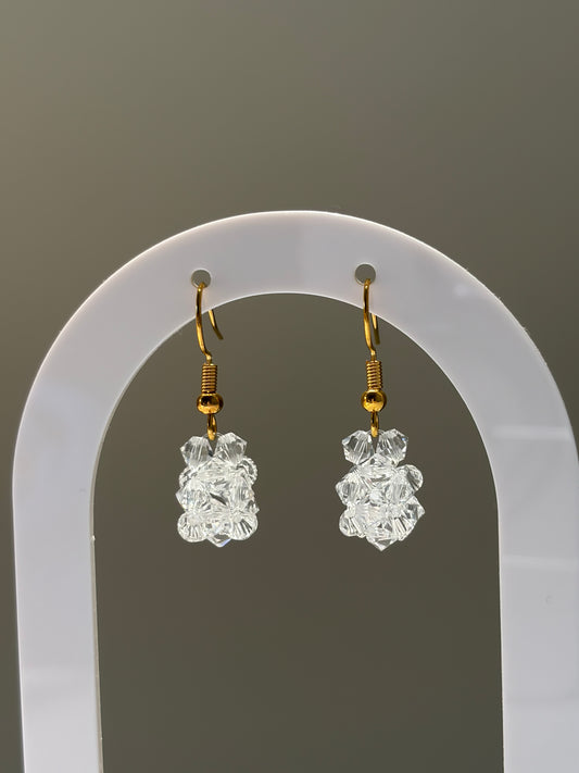 A pair of gold-tone dangle earrings with clear faceted Swarovski crystal beads, displayed on a white arched jewelry stand against a neutral gray background.
