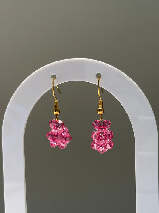 A pair of gold-tone dangle earrings with clustered pink faceted Swarovski crystal beads, displayed on a white arched stand against a neutral gray background.
