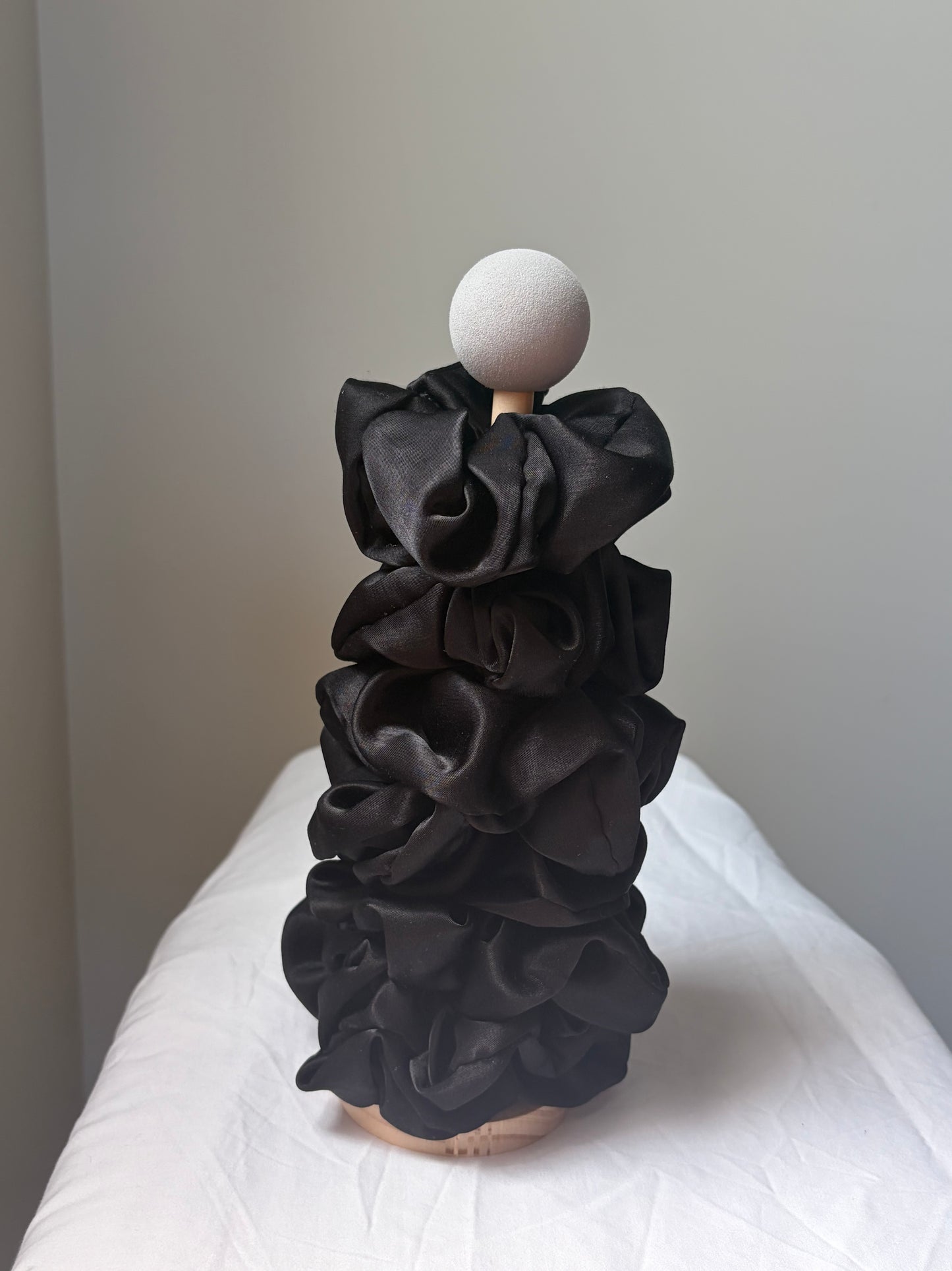 A black silk fabric scrunchies displayed on a mannequin with a round white head and wooden neck, set against a plain gray wall and white draped fabric backdrop.