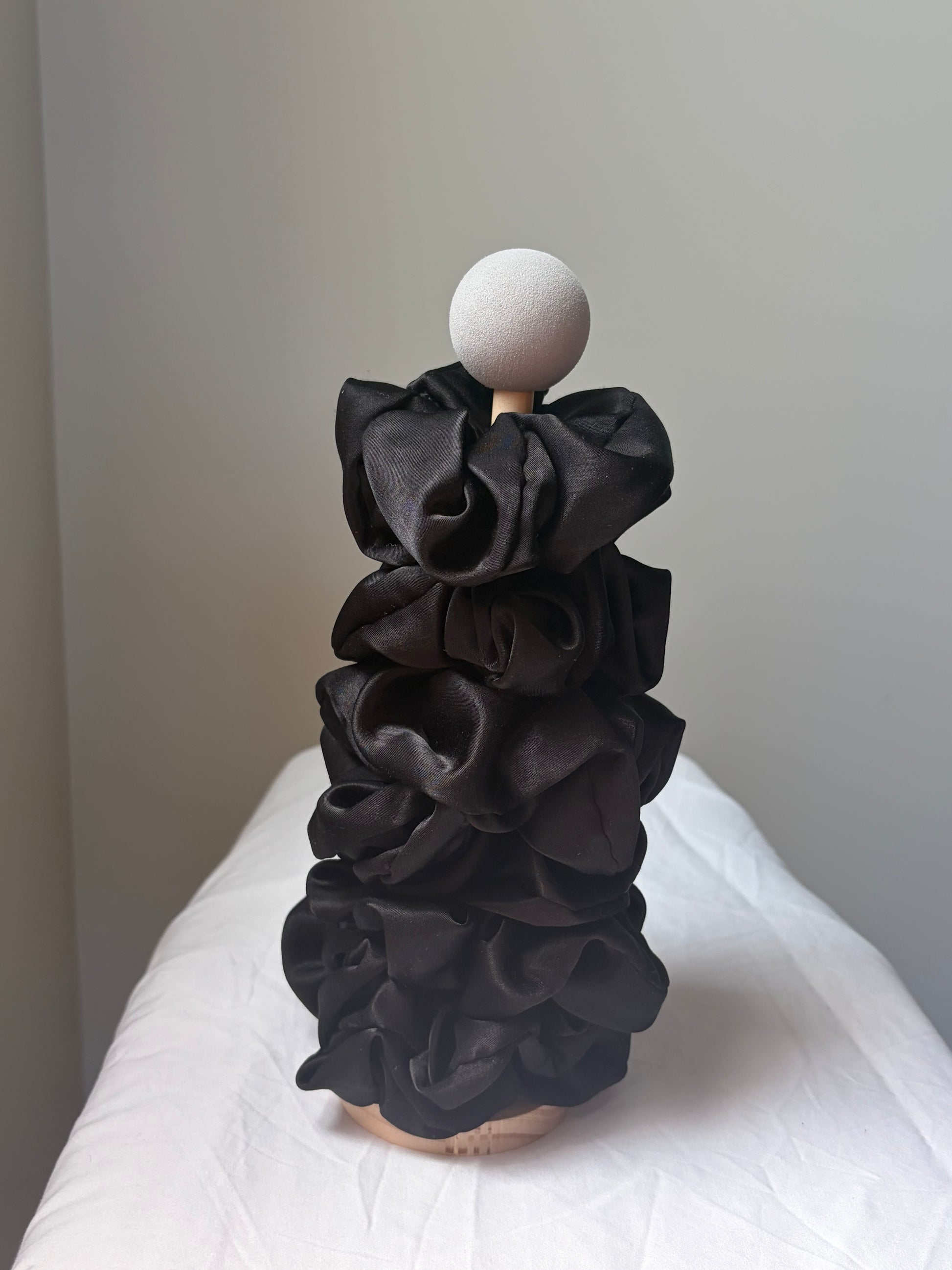 A black silk fabric scrunchies displayed on a mannequin with a round white head and wooden neck, set against a plain gray wall and white draped fabric backdrop.