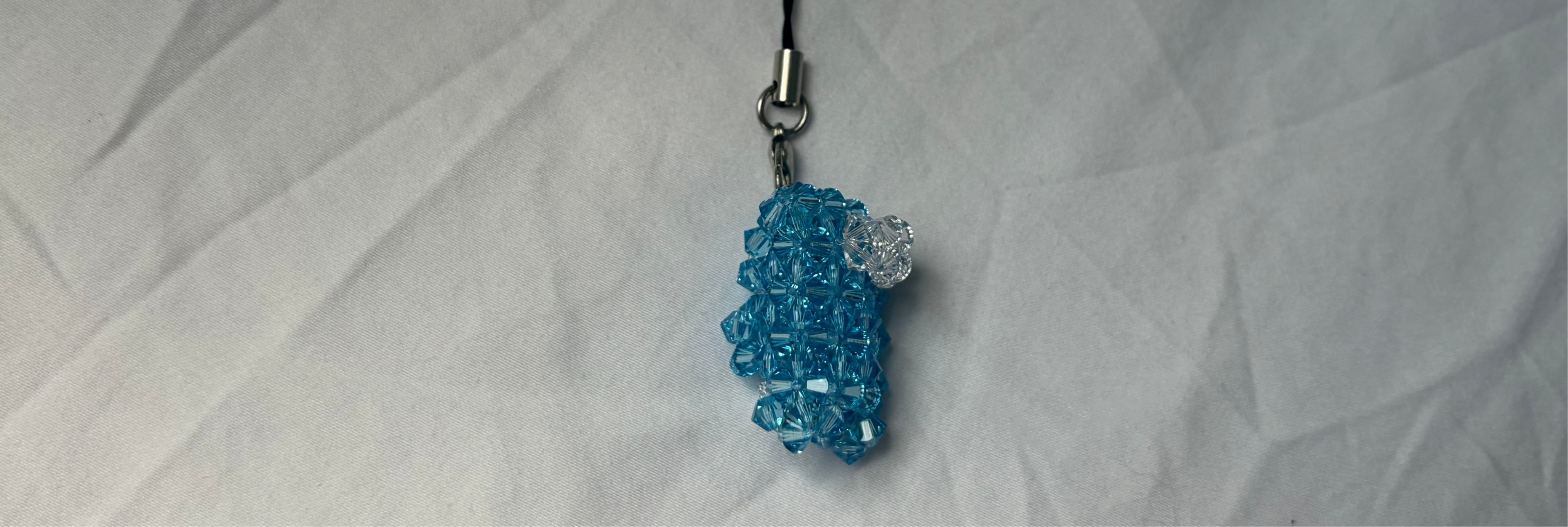 A handmade Among Us beaded charm featuring blue Swarovski crystal beads, a small silver metal clasp, and a clear faceted crystal accent, attached to a black elastic loop strap, displayed on a white fabric background.