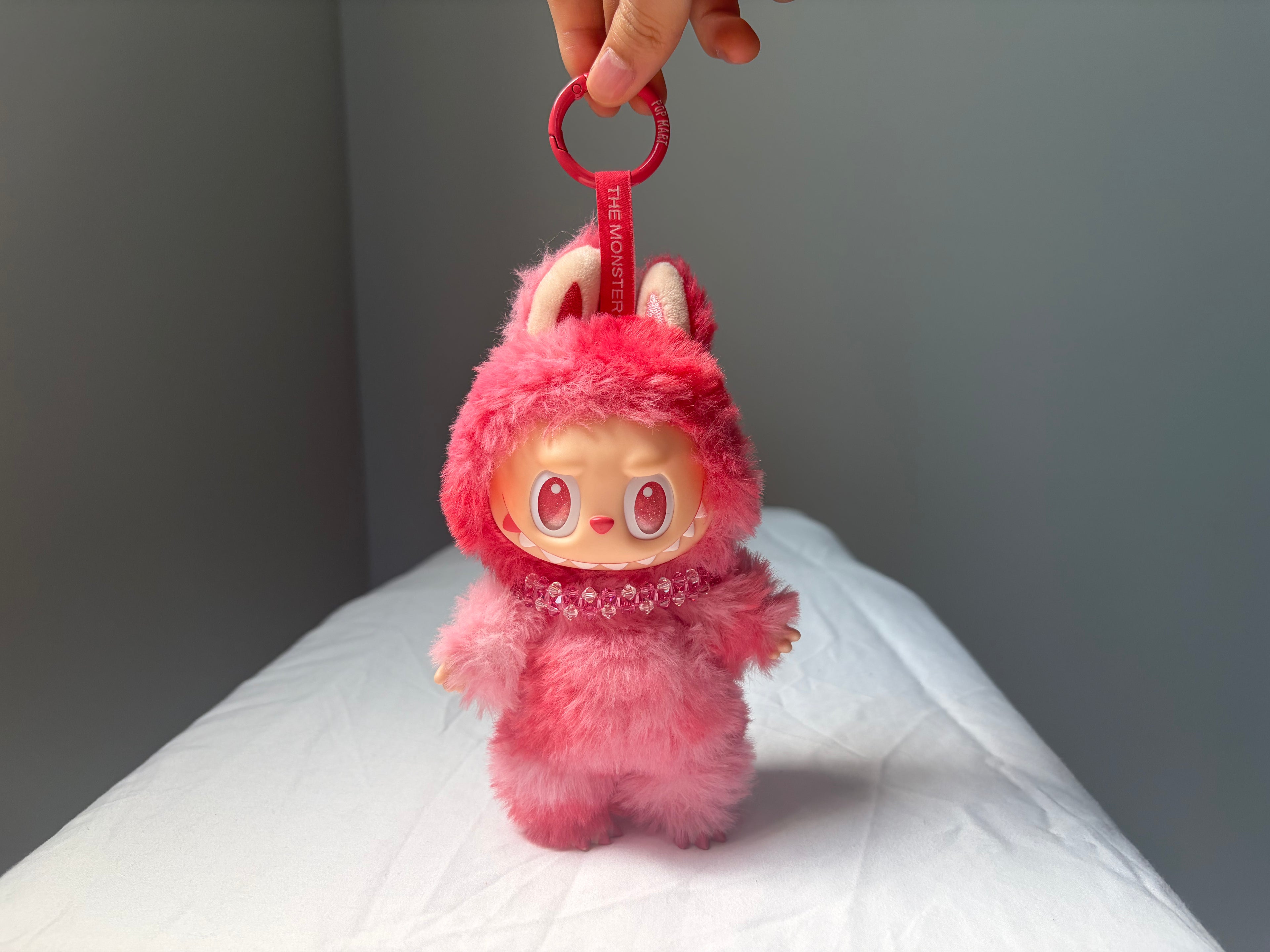 A pink fuzzy Labubu character keychain doll with a red key ring and strap, held by a hand over a white surface against a gray background.