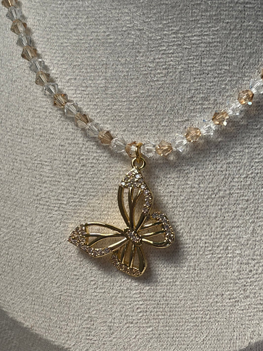 A gold butterfly pendant necklace with a beaded chain of clear and champagne-colored Swarovski crystal faceted beads, displayed on a textured beige fabric bust.