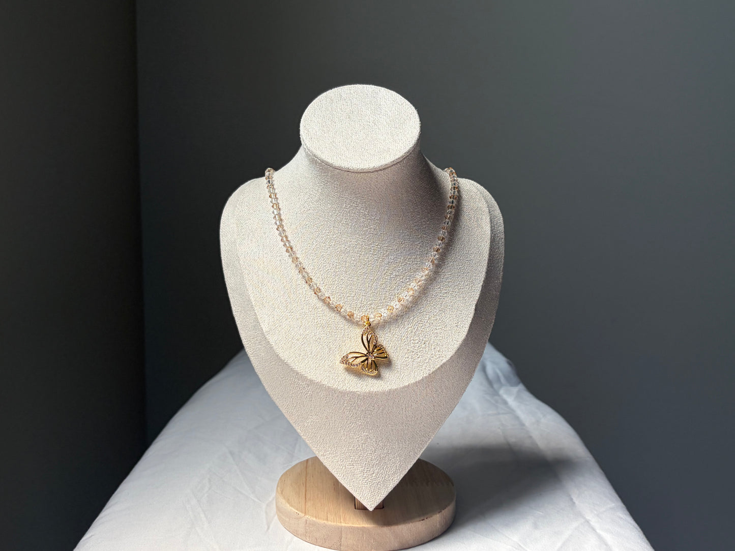 A delicate necklace displayed on a textured cream jewelry bust. The necklace features a single strand of small, gold and white Swarovski crystal beads with a polished gold butterfly pendant at the center.