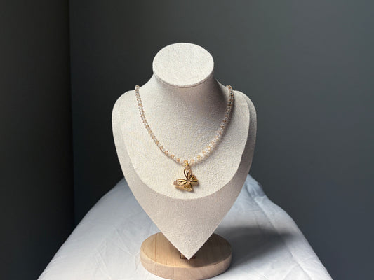 A delicate necklace displayed on a textured cream jewelry bust. The necklace features a single strand of small, gold and white Swarovski crystal beads with a polished gold butterfly pendant at the center.