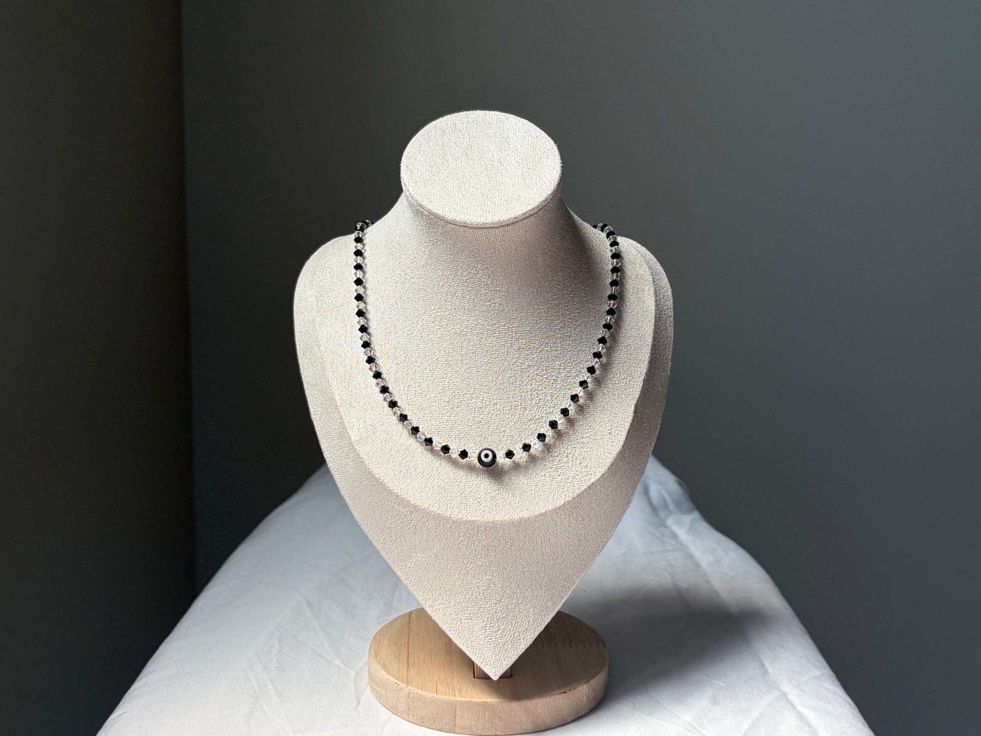A Swarovski crystal beaded necklace with black and white evil eye beads displayed on a textured cream-colored jewelry bust against a dark gray background.