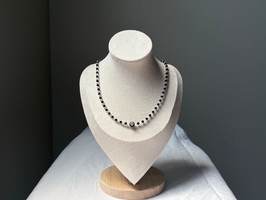 A Swarovski crystal beaded necklace with black and white evil eye beads displayed on a textured cream-colored jewelry bust against a dark gray background.