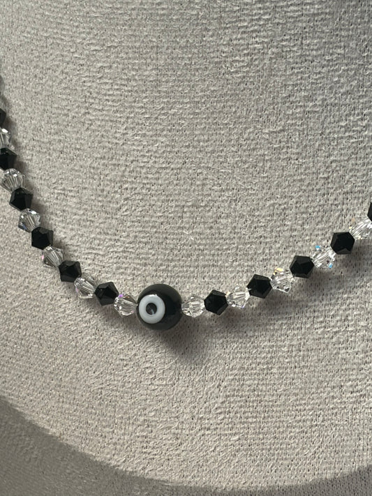 A Swarovski crystal beaded necklace displayed on a textured gray jewelry bust, featuring black faceted beads, clear crystal beads, and a central black evil eye charm with a white and black pupil.