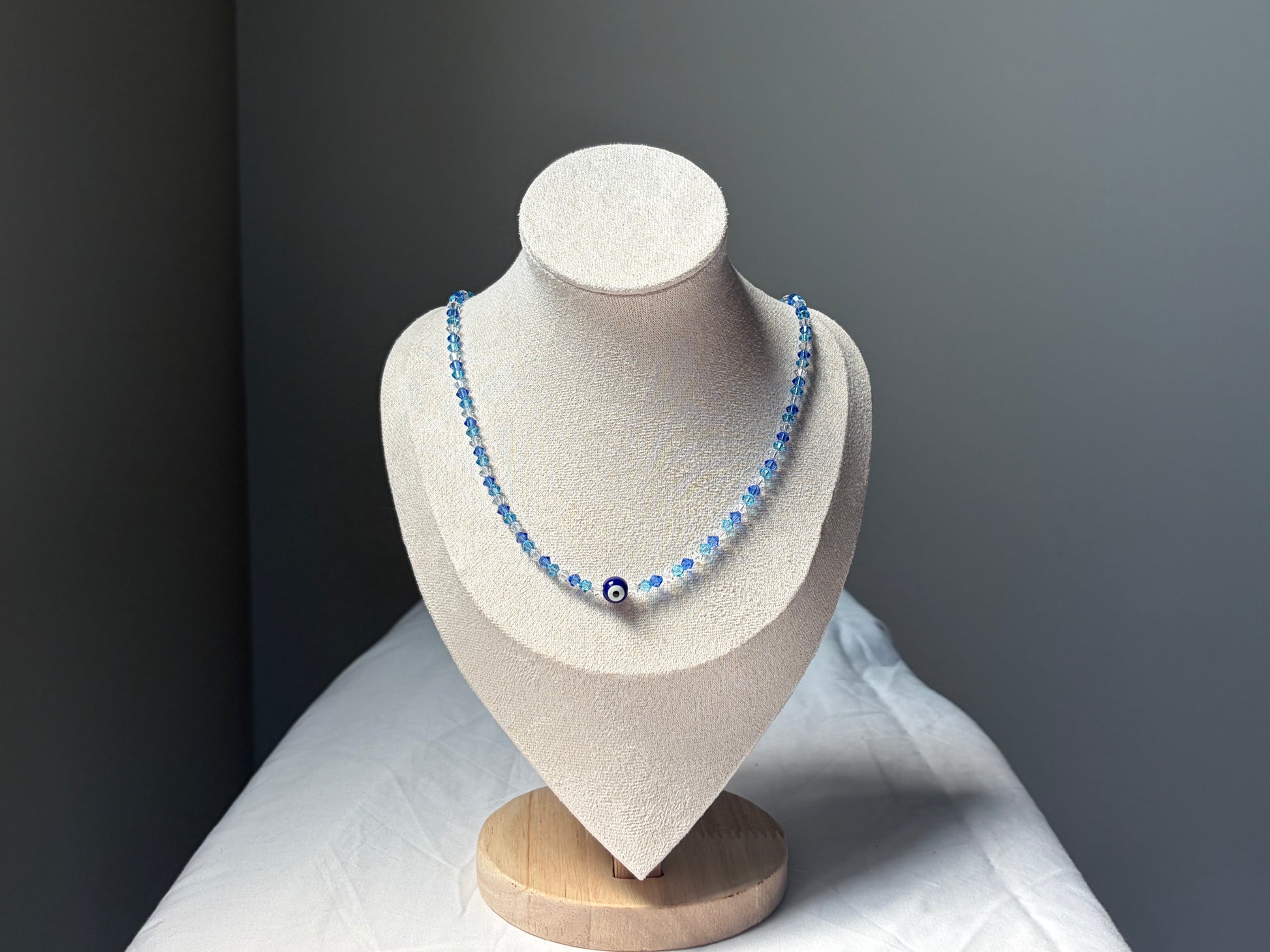 A Swarovski crystal beaded necklace with light blue and dark blue beads, featuring a central evil eye charm, displayed on a textured beige jewelry bust against a dark gray background.