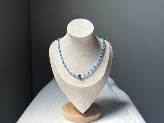 A Swarovski crystal beaded necklace with light blue and dark blue beads, featuring a central evil eye charm, displayed on a textured beige jewelry bust against a dark gray background.