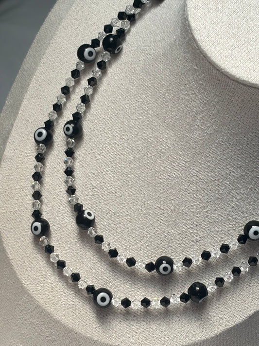 A double-strand Swarovski crystal beaded necklace displayed on a textured cream jewelry bust. The necklace features alternating strands of faceted black beads, clear crystal beads, and round black evil eye charms with white centers.