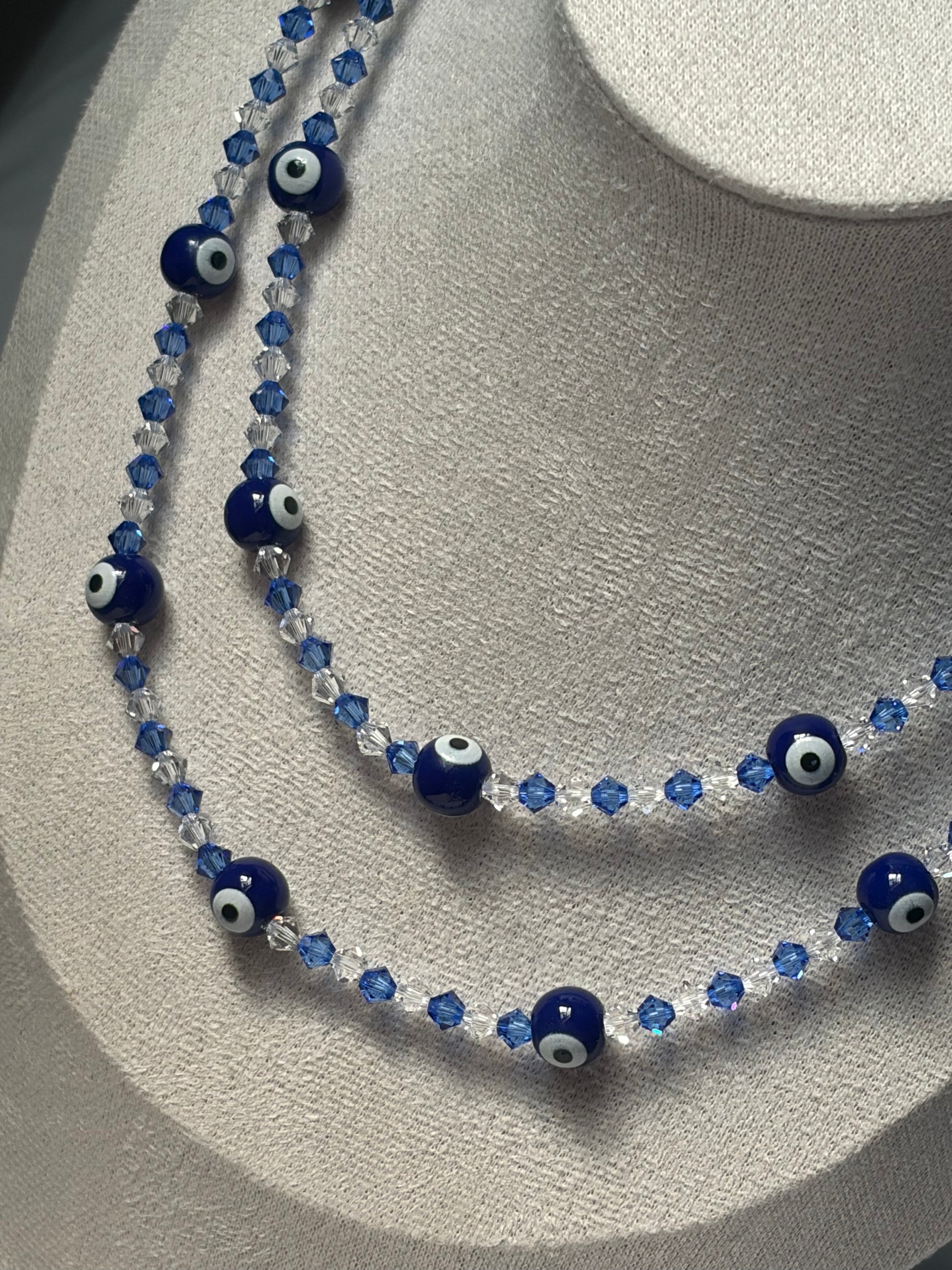 A double-strand Swarovski beaded necklace displayed on a textured light gray jewelry bust. The necklace features dark blue glass evil eye beads with white and black centers, alternating with faceted blue and clear crystal beads.