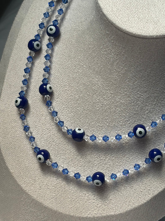 A double-strand Swarovski beaded necklace displayed on a textured light gray jewelry bust. The necklace features dark blue glass evil eye beads with white and black centers, alternating with faceted blue and clear crystal beads.