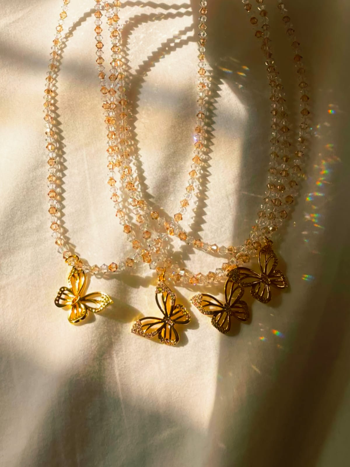 Three Swarovski crystal beaded necklaces with gold butterfly pendants draped on a cream fabric background with warm sunlight and shadow patterns.