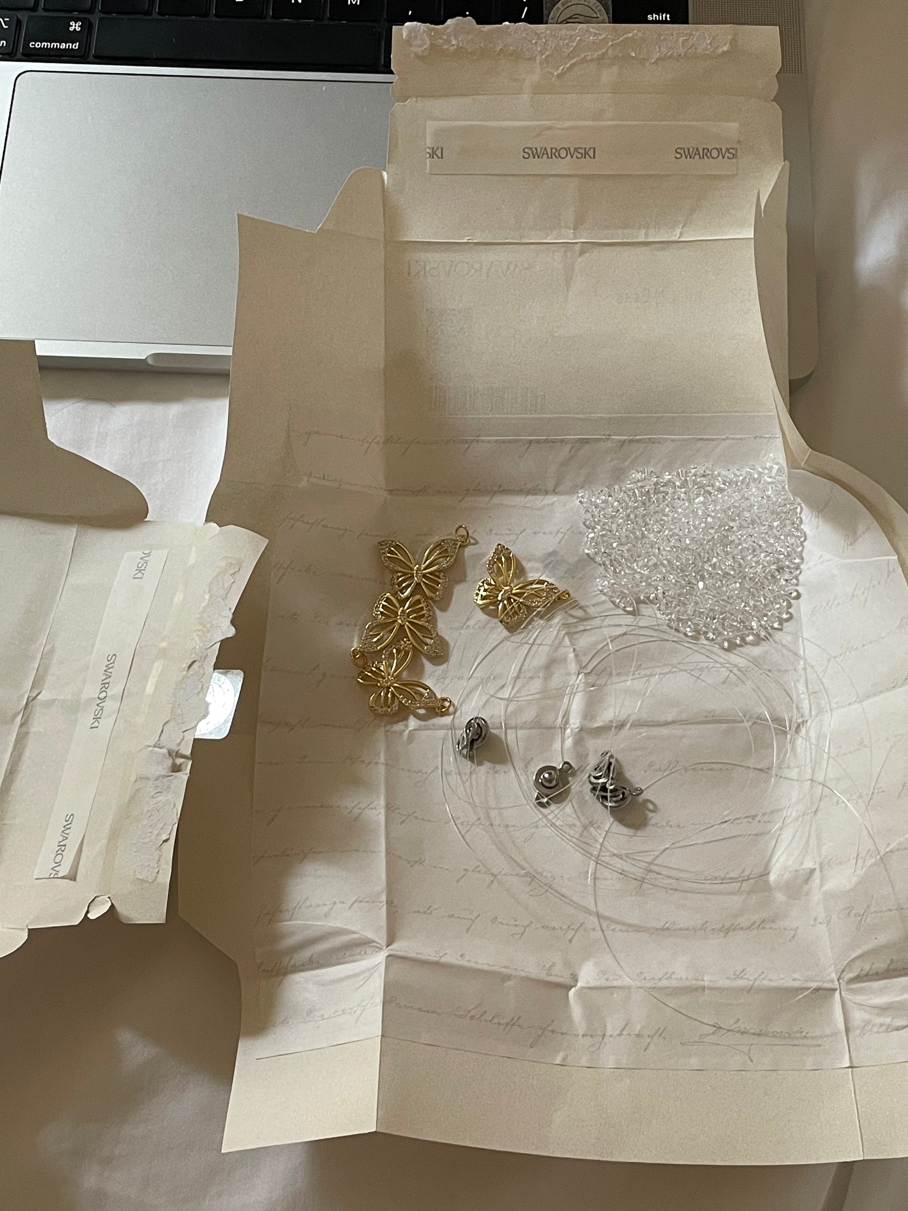 Jewelry components laid out on a white Swarovski branded paper insert, including gold butterfly-shaped charms, silver and clear Swarovski crystal beads, and small stainless steel jewelry clasps, with a laptop partially visible in the background.