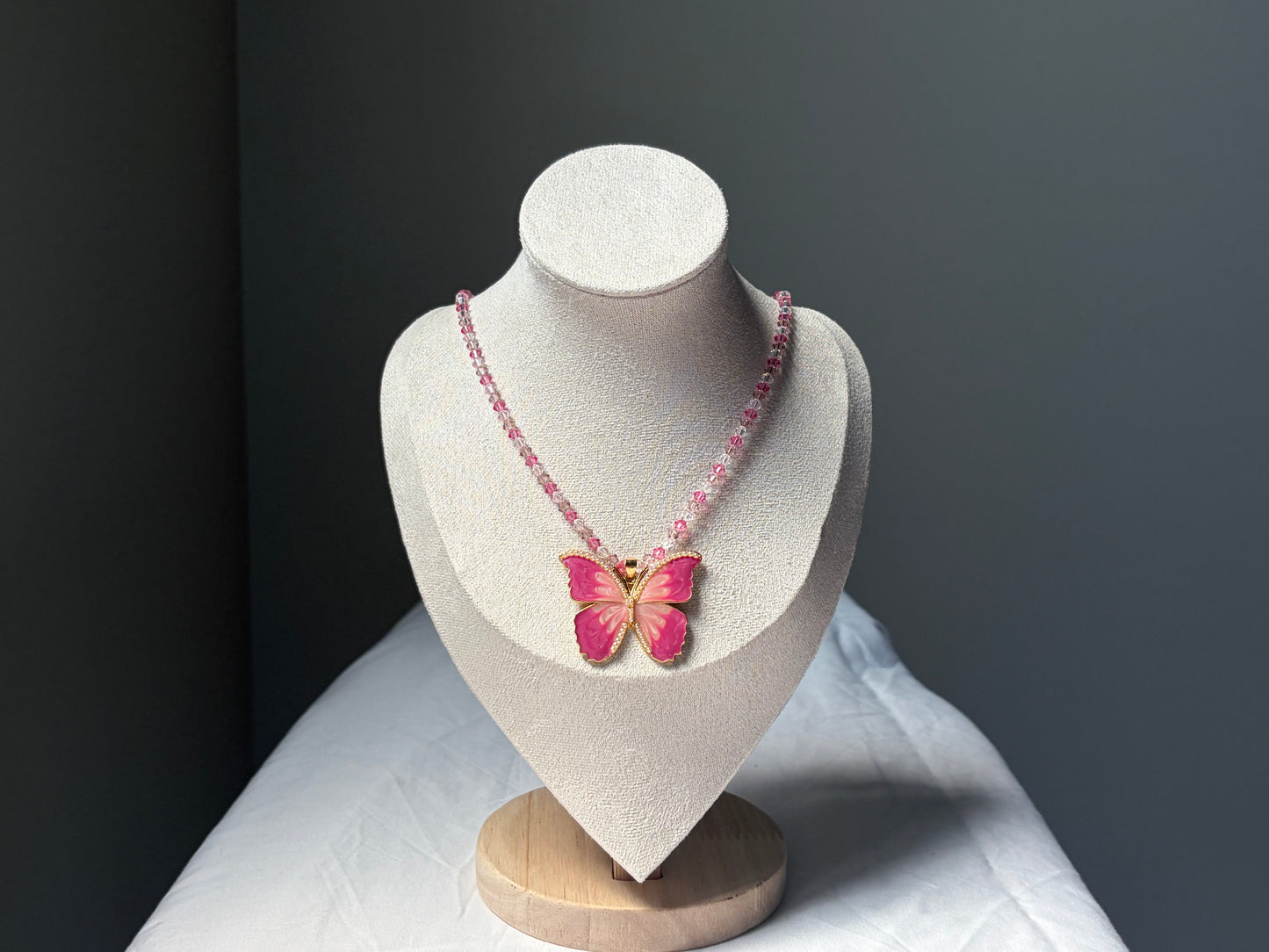 A pink Swarovski crystal beaded necklace with a butterfly pendant displayed on a textured white jewelry bust against a dark gray background.