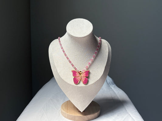 A pink Swarovski crystal beaded necklace with a butterfly pendant displayed on a textured white jewelry bust against a dark gray background.