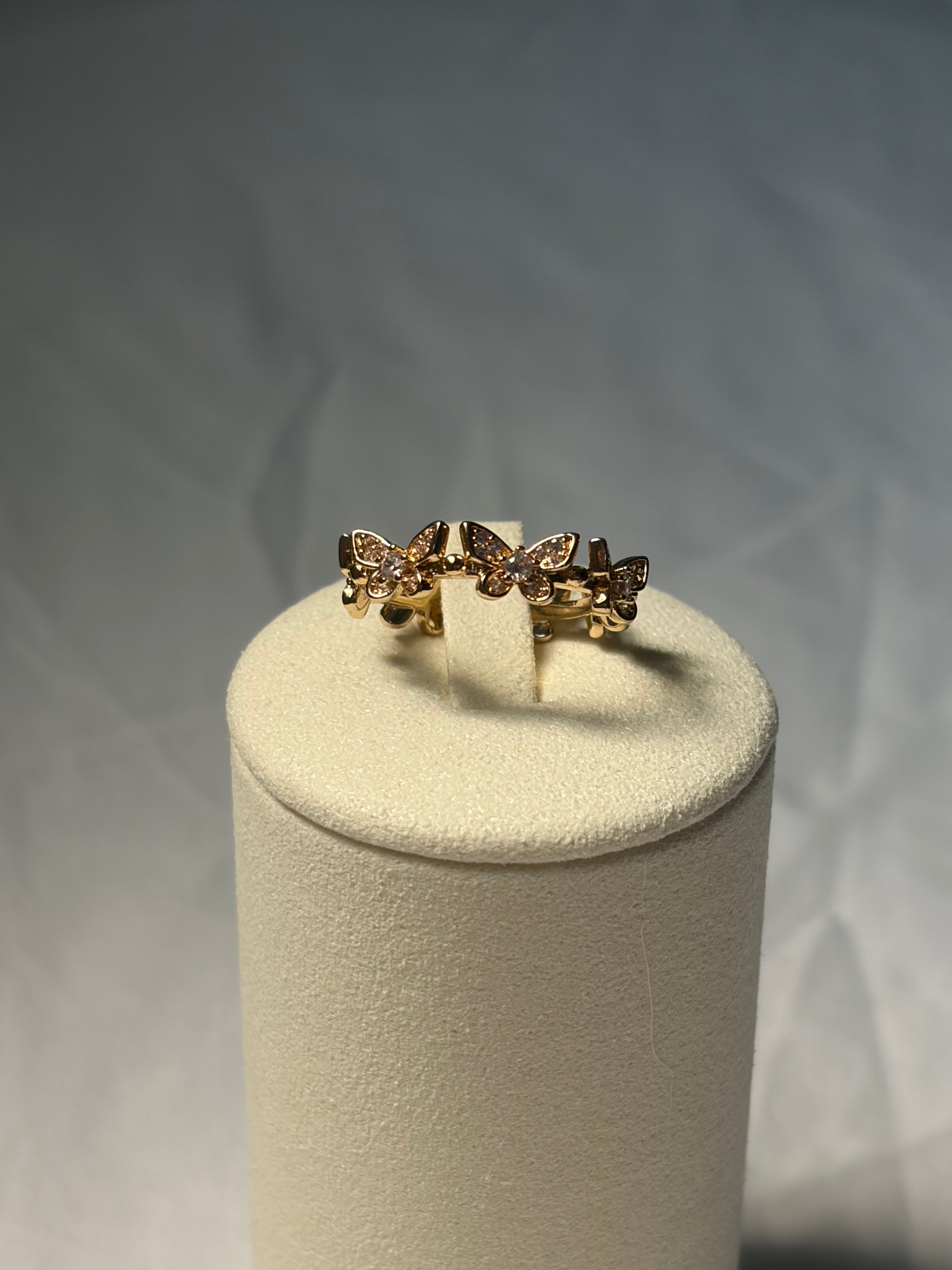 A gold ring with three butterfly-shaped settings, each holding small clear stones, displayed on a cream-colored cylindrical jewelry stand against a gray background.