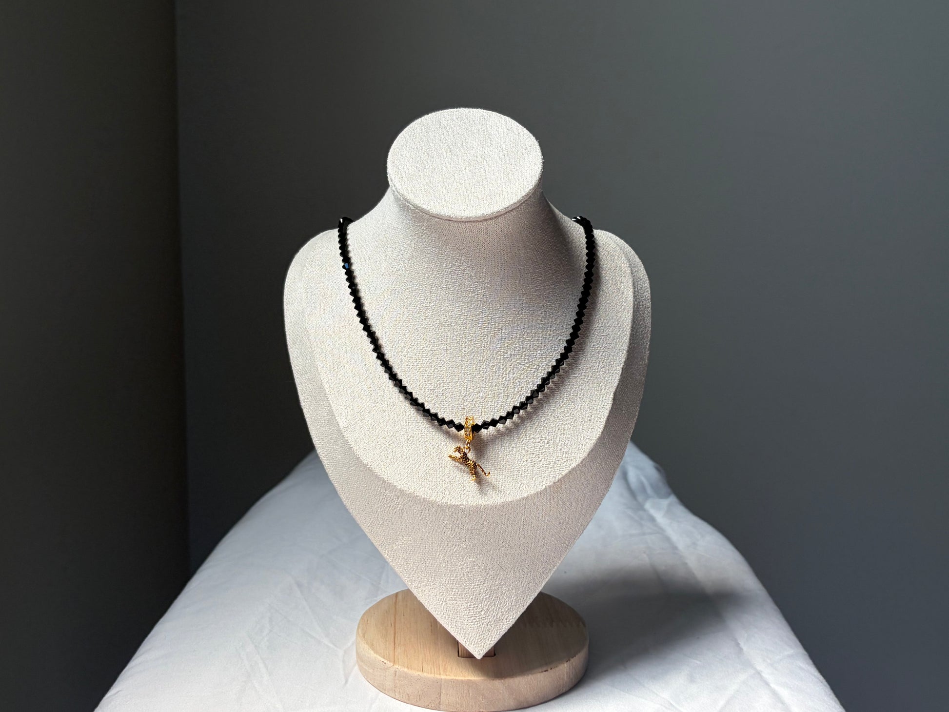 A black Swarovski crystal beaded necklace with a small gold-toned leopard charm pendant displayed on a white jewelry bust against a dark gray background.