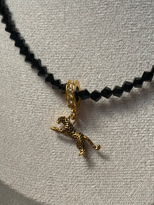 A close-up of a necklace with a black faceted Swarovski crystal bead chain and a gold-toned leopard pendant, displayed on a textured light beige surface.