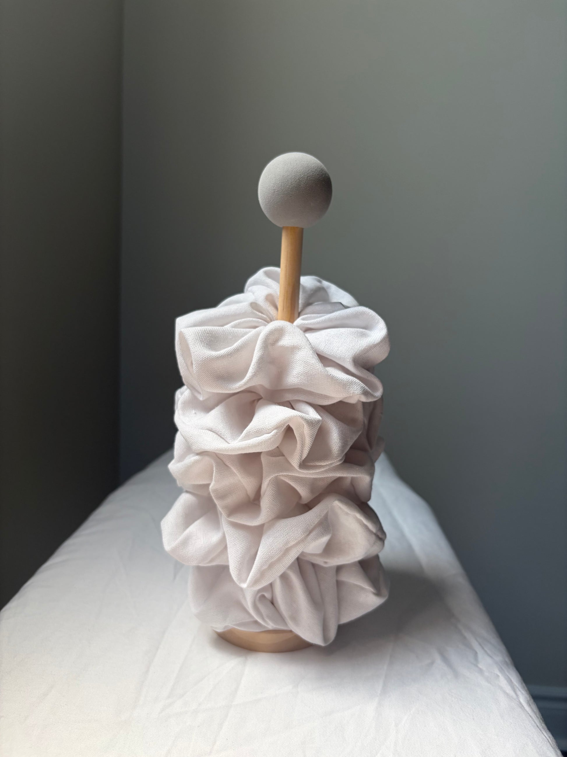 A decorative sculptural object with five layered ruffled linen scrunchie, mounted on a light wood stand with a round wooden top, photographed indoors against a dark gray wall and white draped surface.