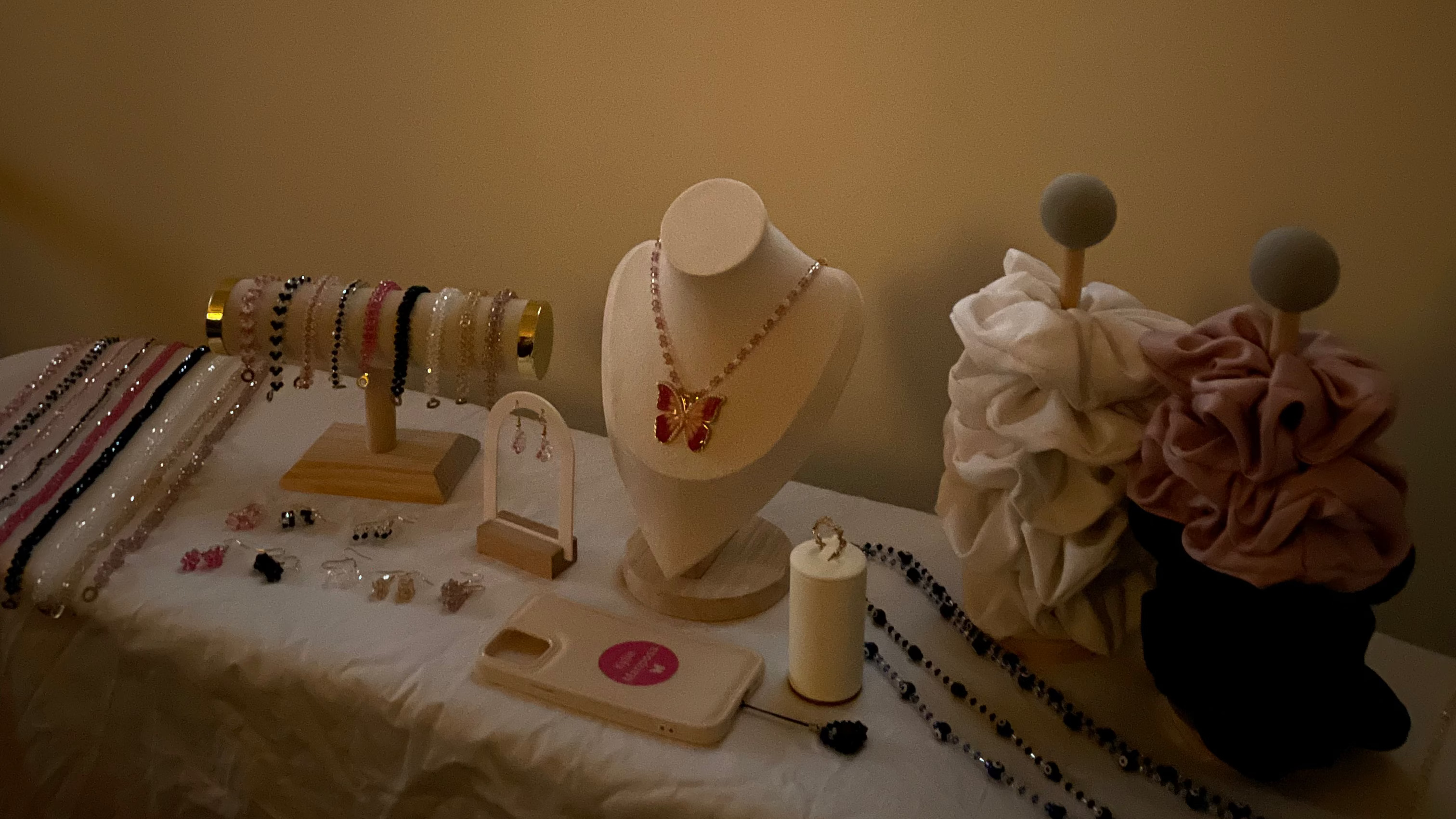 A styled jewelry display on a white-covered table beneath a window, featuring necklaces on a mannequin bust, earrings on stands, draped fabric jewelry holders, and beads. 