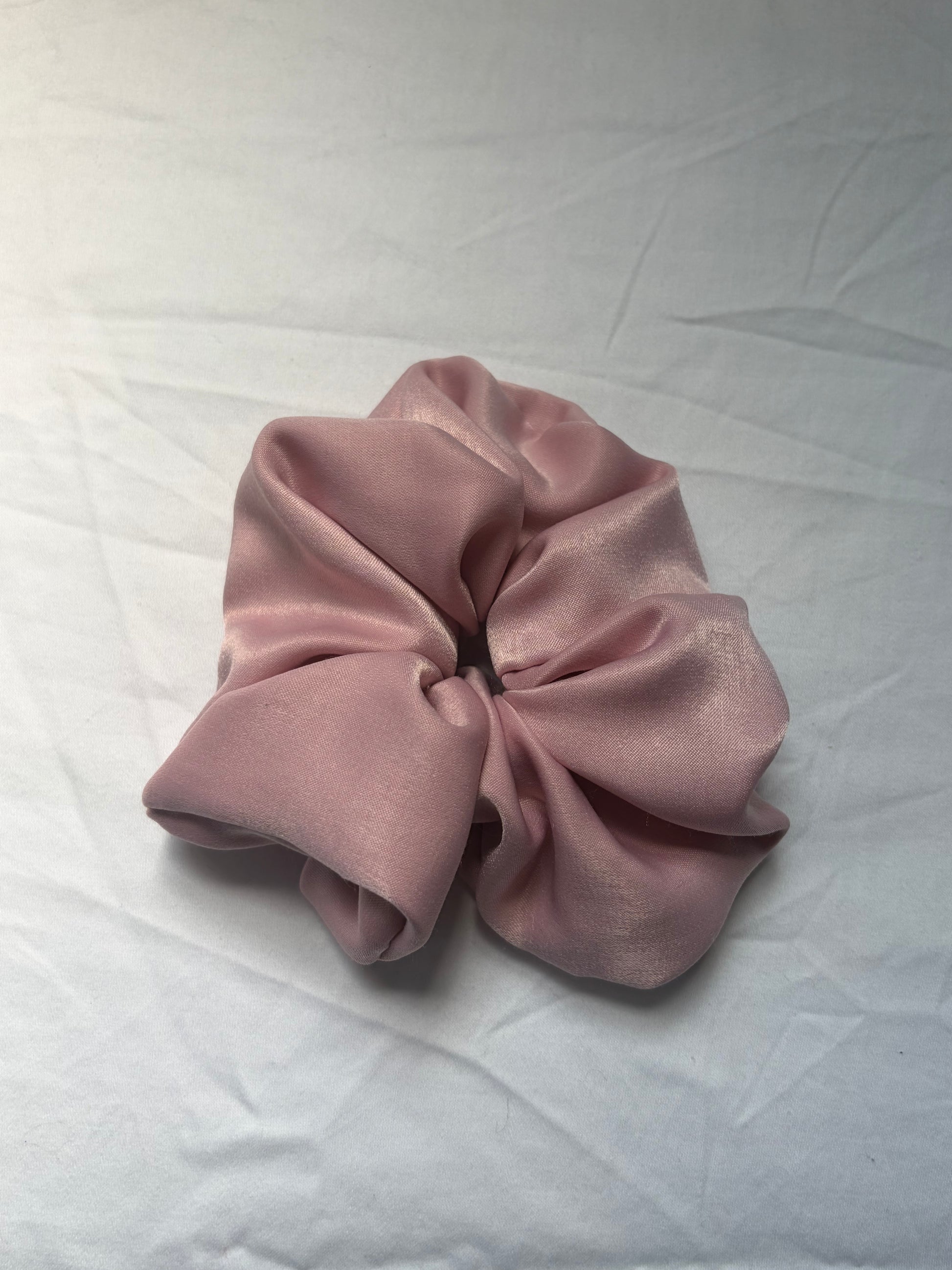 A dusty pink satin hair scrunchie resting on a white fabric surface.