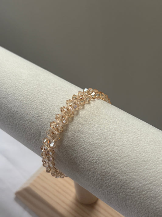 A delicate gold-tone bracelet with clear faceted Swarovski crystal beads, displayed on a textured cream jewelry stand.