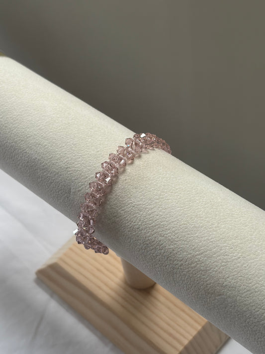 A delicate rose gold pink Swarovski crystal beaded bracelet displayed on a cream-colored jewelry stand with a light wood base against a neutral gray background.