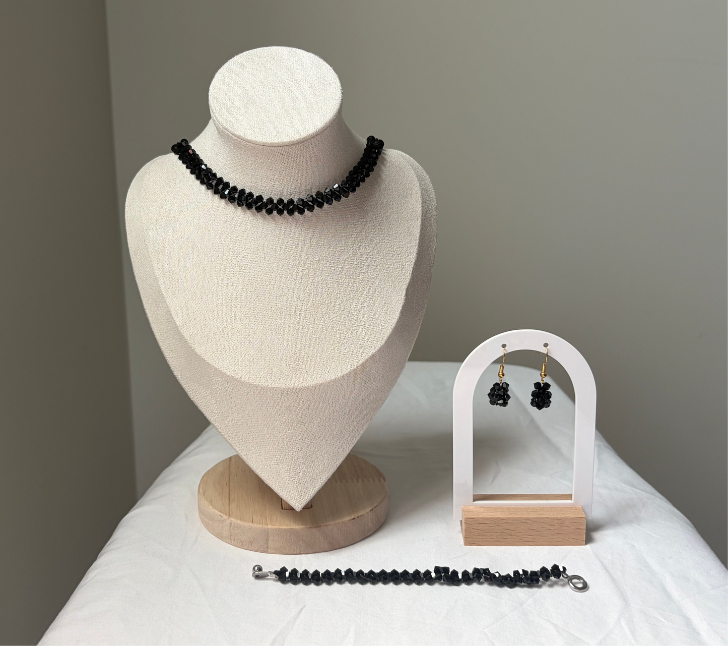 A Swarovski crystal beaded jewelry set displayed on a cream fabric mannequin bust and a small white arch stand, featuring a necklace, bracelet, and matching earrings with black solid design. 