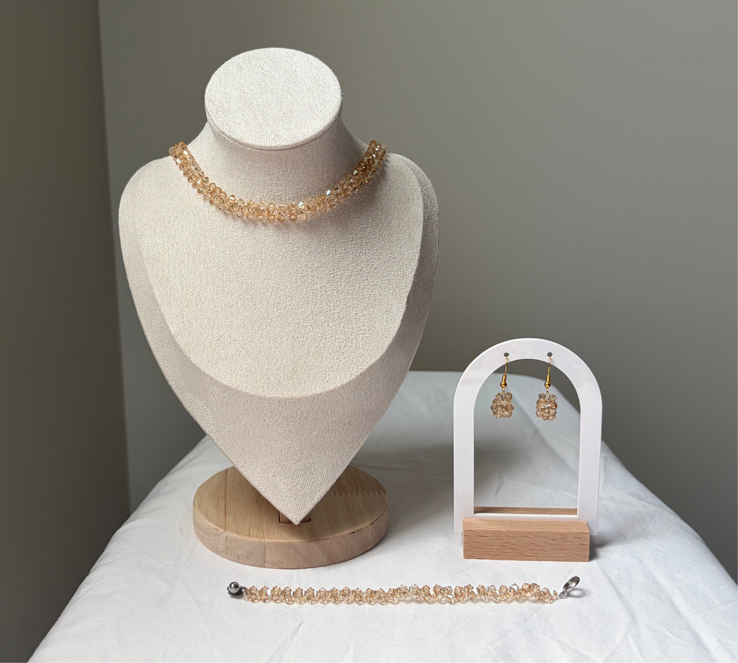 A Swarovski crystal beaded jewelry set displayed on a cream fabric mannequin bust and a small white arch stand, featuring a necklace, bracelet, and matching earrings with champagne gold solid design. 