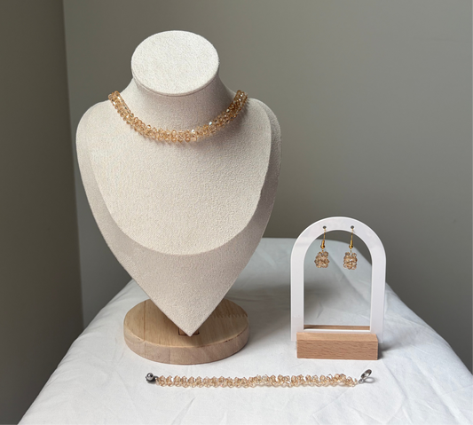 A Swarovski crystal beaded jewelry set displayed on a cream fabric mannequin bust and a small white arch stand, featuring a necklace, bracelet, and matching earrings with champagne gold solid design. 