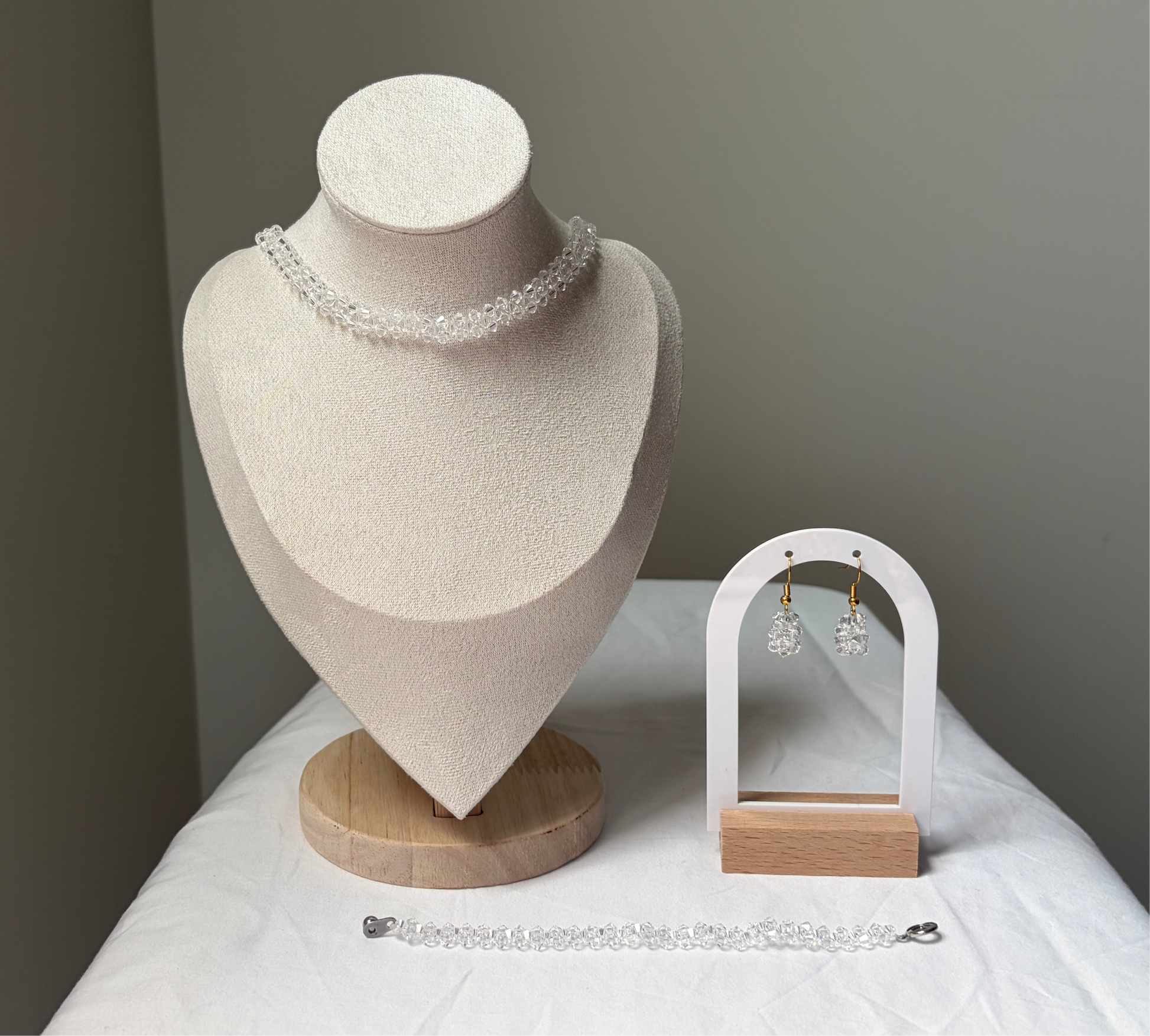 A Swarovski crystal beaded jewelry set displayed on a cream fabric mannequin bust and a small white arch stand, featuring a necklace, bracelet, and matching earrings with white clear solid design. 