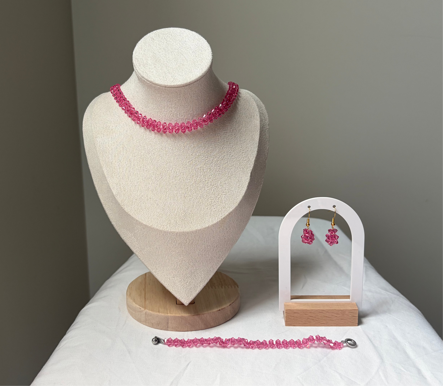 A Swarovski crystal beaded jewelry set displayed on a cream fabric mannequin bust and a small white arch stand, featuring a necklace, bracelet, and matching earrings with pink solid design. 