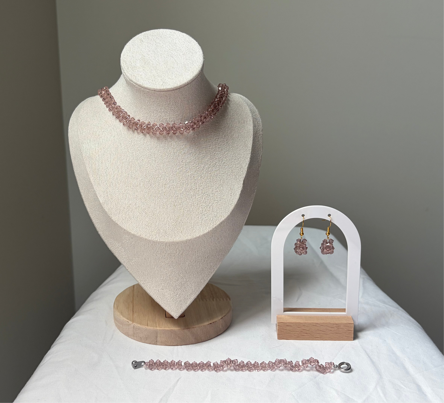 A Swarovski crystal beaded jewelry set displayed on a cream fabric mannequin bust and a small white arch stand, featuring a necklace, bracelet, and matching earrings with pink rose gold solid design. 