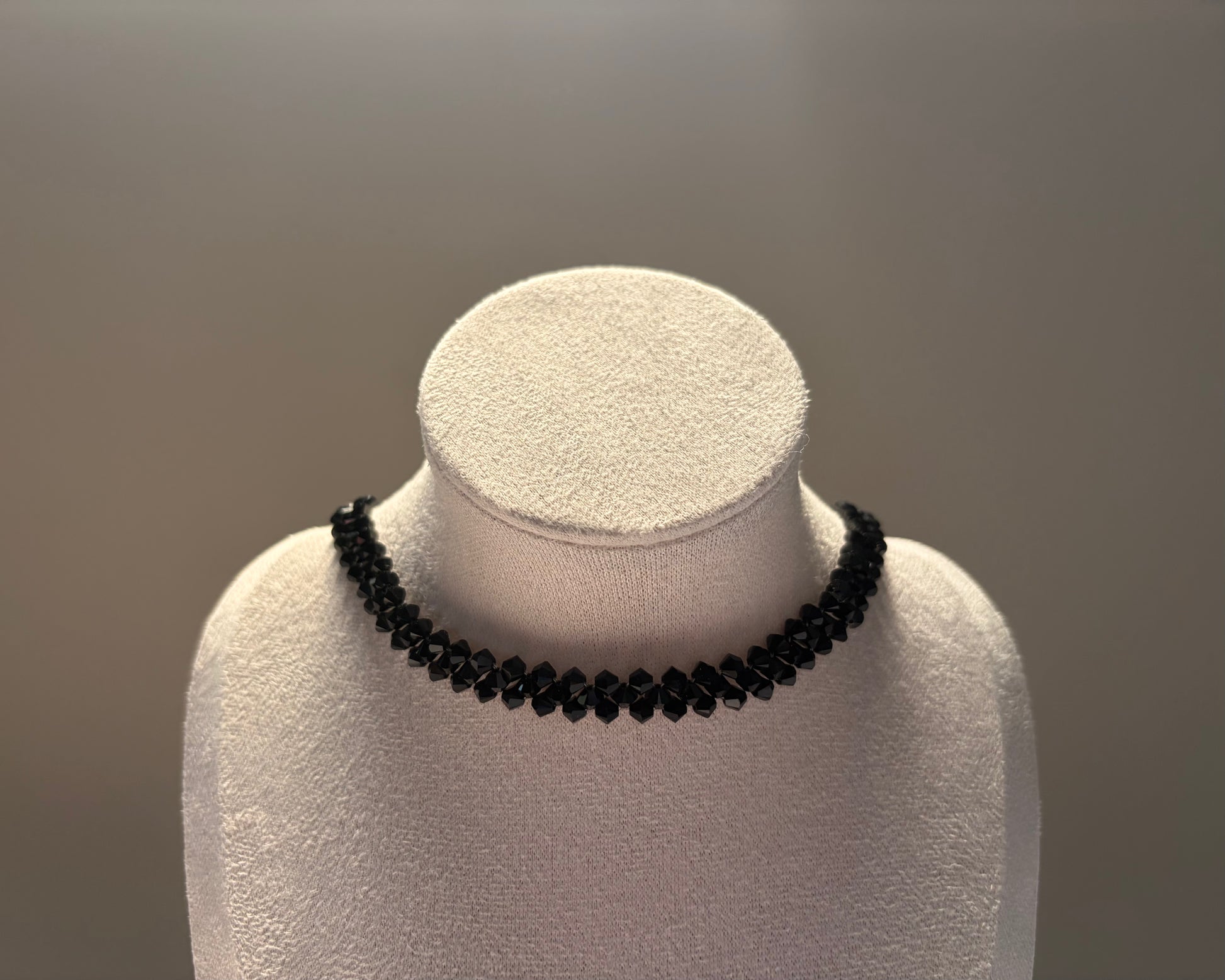 A black Swarovski crystal beaded solid choker necklace displayed on a white textured jewelry bust against a neutral gray background.