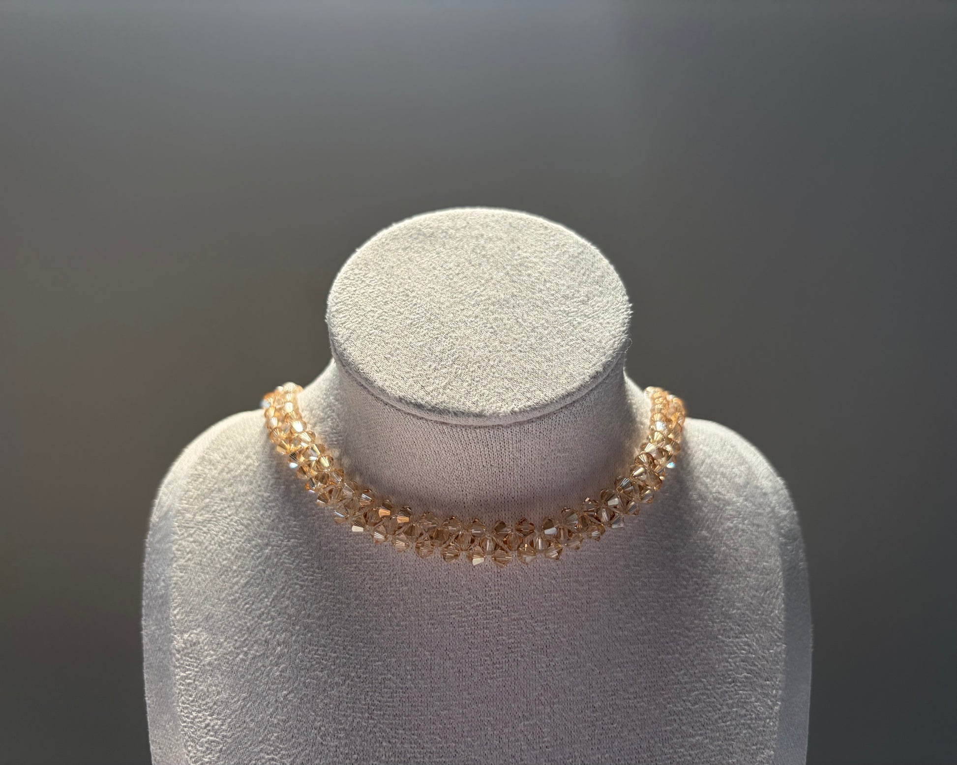 A delicate gold-toned Swarovski crystal beaded solid choker necklace displayed on a textured light gray jewelry bust against a dark gray gradient background.