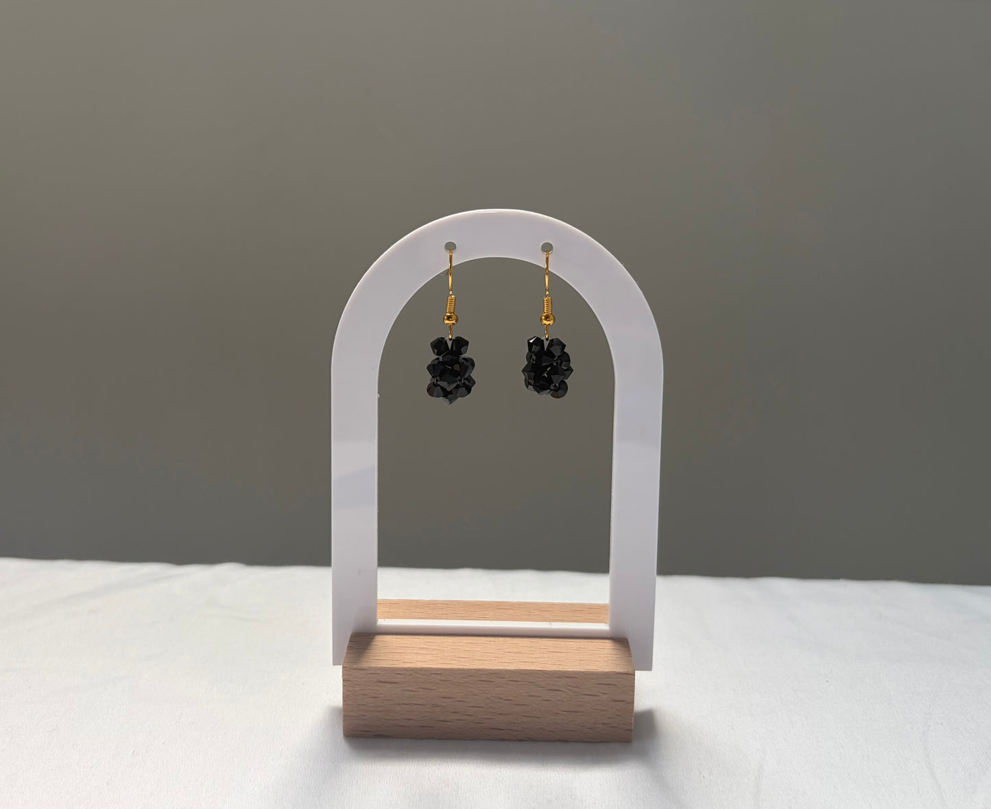 A pair of black solid shaped Swarovski crystal beaded dangle earrings with gold hooks, displayed on a white arch stand with a wooden base against a neutral gray background.