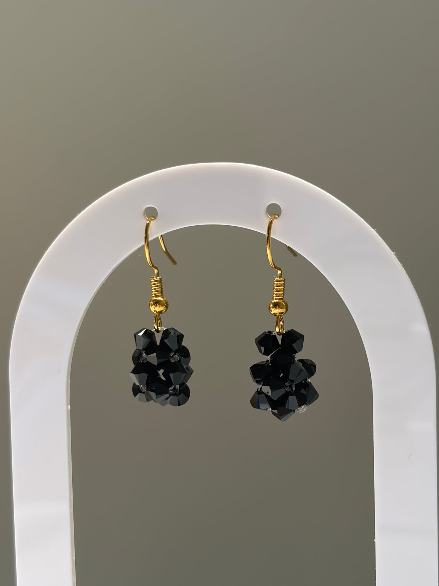 A pair of gold-tone dangle earrings with faceted black Swarovski crystal beaded clusters, displayed on a white arched stand against a neutral gray background.
