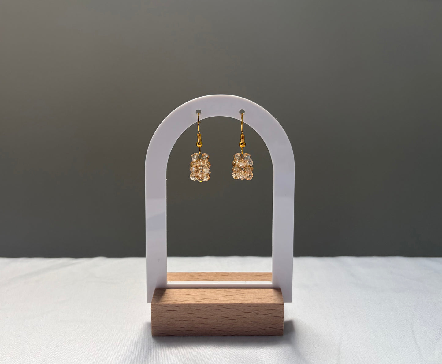 A pair of gold-tone drop earrings with textured, solid shaped gold-toned Swarovski crystal beaded material, displayed on a white arch stand with a wooden base against a dark gray background.