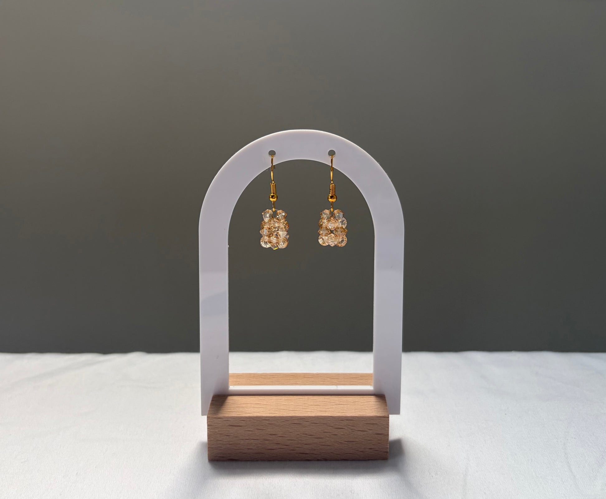 A pair of gold-tone drop earrings with textured, solid shaped gold-toned Swarovski crystal beaded material, displayed on a white arch stand with a wooden base against a dark gray background.