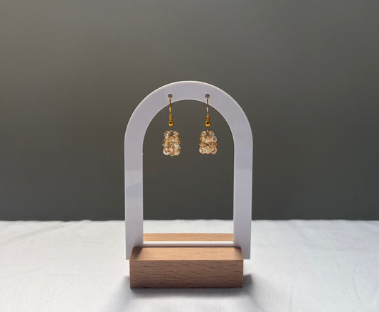 A pair of gold-tone drop earrings with textured, solid shaped gold-toned Swarovski crystal beaded material, displayed on a white arch stand with a wooden base against a dark gray background.