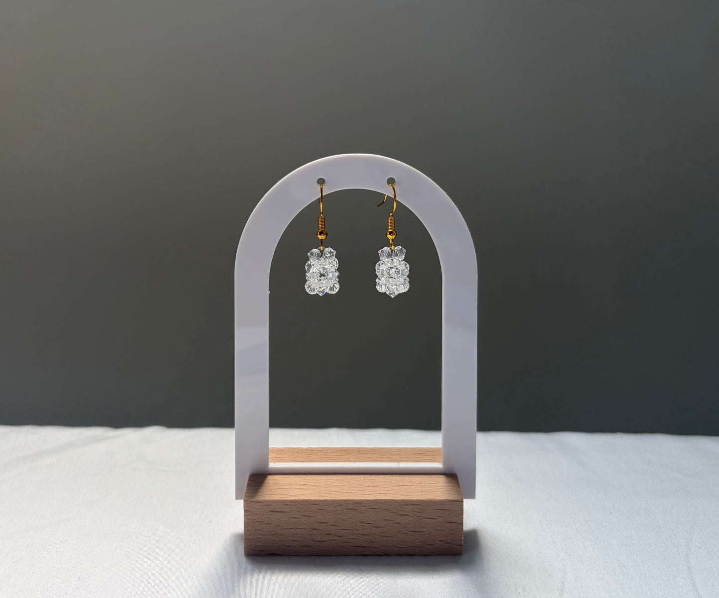 A pair of small clear Swarovski crystal beaded dangle earrings with gold hooks, displayed on a white arch-shaped stand with a wooden base against a dark gray background.