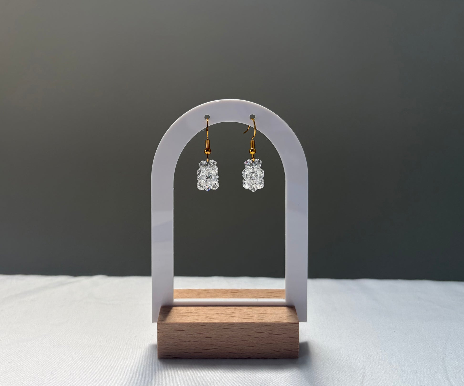 A pair of small clear Swarovski crystal beaded dangle earrings with gold hooks, displayed on a white arch-shaped stand with a wooden base against a dark gray background.