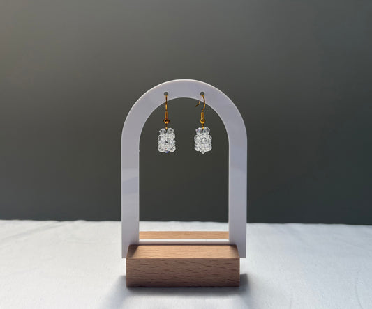 A pair of small clear Swarovski crystal beaded dangle earrings with gold hooks, displayed on a white arch-shaped stand with a wooden base against a dark gray background.